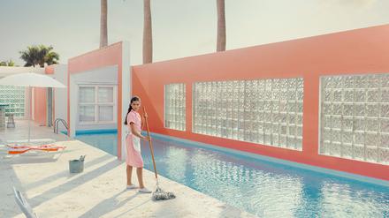 Das Foto „Pink Dreams #2, Miami Shores“ von Dean West zeigt eine Reinigungsfrau, die mit einem Wischmopp in der Hand neben einem blau schimmerndem Pool steht. 