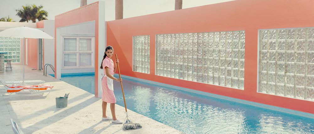 Das Foto „Pink Dreams #2, Miami Shores“ von Dean West zeigt eine Reinigungsfrau, die mit einem Wischmopp in der Hand neben einem blau schimmerndem Pool steht.
