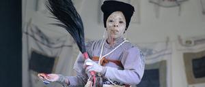 Eine Frau mit einem weiß geschminkten Gesicht in historischer Arbeitskleidung einer Hausangestellten hält einen Federschweif in der Hand. Es ist die Künstlerin Tuli Mekondjo bei der Perfomance “Ousie Martha”. 