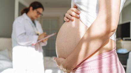 Schwangere Frau zeigt ihren Babybauch, im Hintergrund macht sich eine Ärztin Notizen.