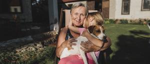 Eine lächelnde Großmutter sitzt mit Enkelin und Hund auf einem Stuhl im Garten.