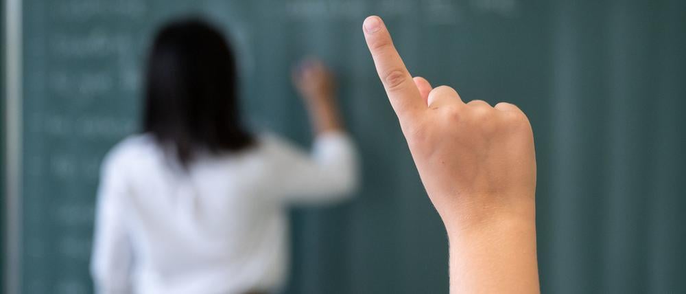 Ein Schüler meldet sich während einer Unterrichtsstunde in einem Gymnasium, während eine Lehrerin an die Tafel schreibt.