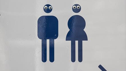 Ein Schild Mann und Frau mit einem Pfeil weist im Park Charlottenburg auf die Toiletten hin. Die Piktogramme sind mit Augen beklebt. 