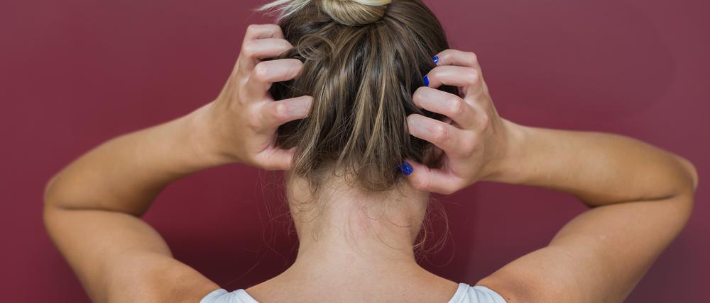 Eine Frau vor rotem Hintergrund fährt sich mit den Händen durch die Haare, um sich zu kratzen.
