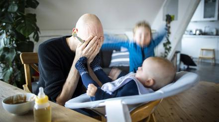 Symbolfoto: Ein Vater haelt sich verzweifelt die Hande vor das Gesicht während er sein Baby füttert und ein weiteres Kind wütend im Hintergrund herumspringt.