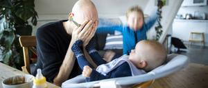 Symbolfoto: Ein Vater haelt sich verzweifelt die Hande vor das Gesicht während er sein Baby füttert und ein weiteres Kind wütend im Hintergrund herumspringt.