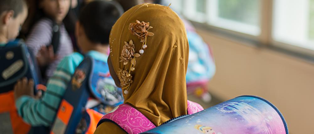 Ein Mädchen mit Kopftuch steht in einer Schule vor einem Klassenzimmer. 