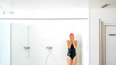 Eine Frau im Badeanzug steht in einer Fitnessstudio-Dusche und hält sich die Augen zu.