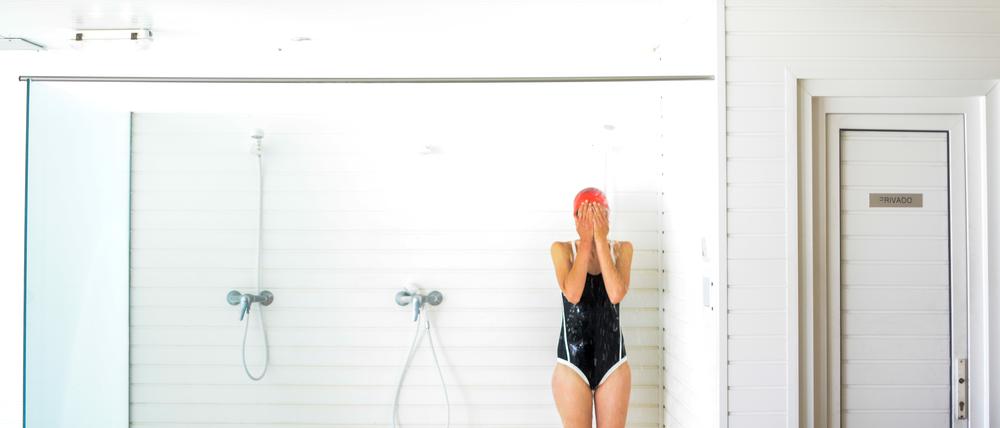 Eine Frau im Badeanzug steht in einer Fitnessstudio-Dusche und hält sich die Augen zu.