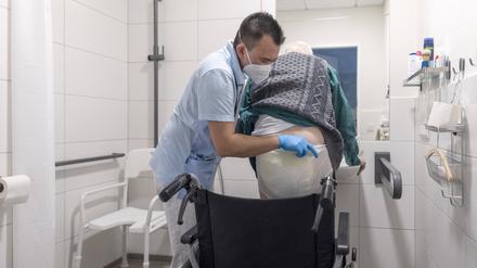 Ein Pfleger hebt einen älteren Mann aus dem Rollstuhl und zieht ihm ein neues Inkontinenzmaterial an, umgangssprachlich für Windel.