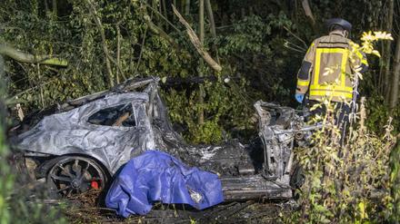 Ein Feuerwehrmann steht im dicht bewachsenen Grünstreifen der Autobahn A44 neben dem Wrack eines ausgebrannten Sportwagens.