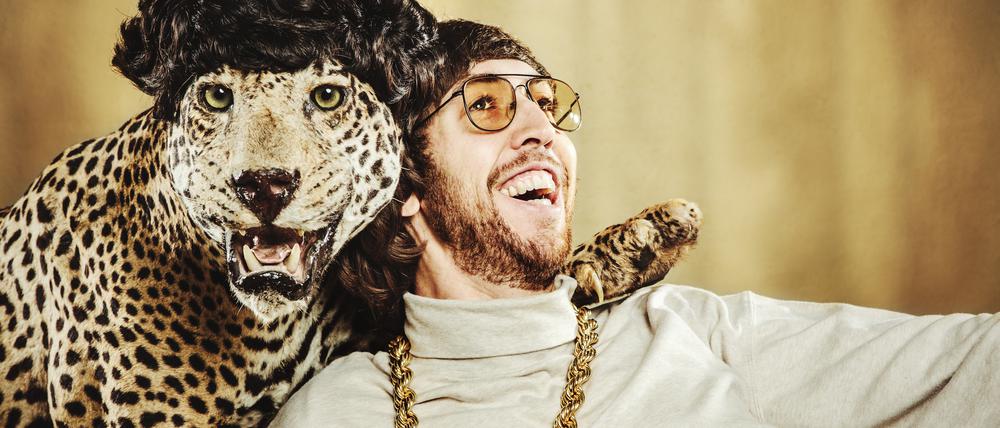 Best friends; a man with shaggy hair and 1970’s style relaxes with a leopard wearing a wig, their arms around each other. 