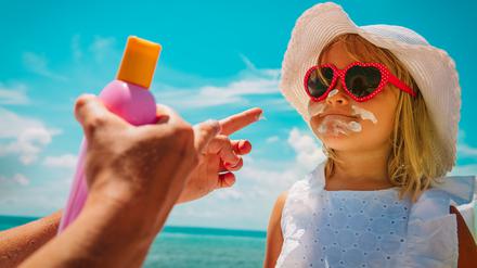 Ein Kind mit Sonnenbrille und Sonnenhut wird gerade mit Sonnencreme eingecremt. Im Vordergrund sind die Hände der Mutter mit der Sonnencremeflasche zu sehen.