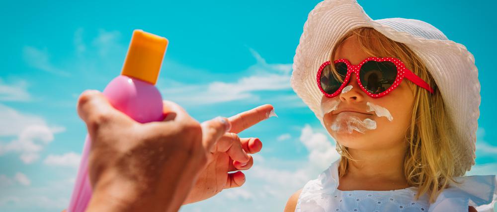Ein Kind mit Sonnenbrille und Sonnenhut wird gerade mit Sonnencreme eingecremt. Im Vordergrund sind die Hände der Mutter mit der Sonnencremeflasche zu sehen.