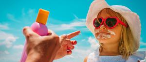 Ein Kind mit Sonnenbrille und Sonnenhut wird gerade mit Sonnencreme eingecremt. Im Vordergrund sind die Hände der Mutter mit der Sonnencremeflasche zu sehen.