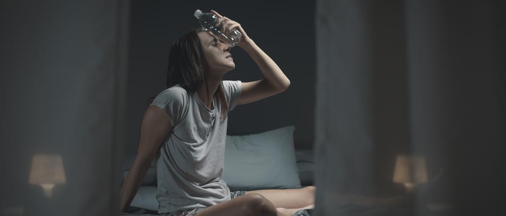 Eine schwitzende Frau auf einem Bett hält sich eine Wasserflasche an die Stirn.