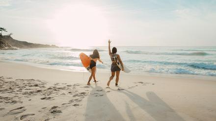 Zwei Menschen mit Surfbrett laufen am Strand.