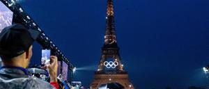Der am Abend beleuchtete Eiffelturm mit olympischen Ringen, davor stehen Zuschauer.