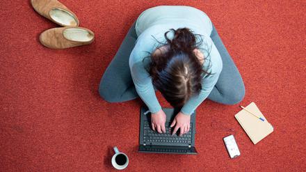 Eine Frau sitzt im Homeoffice auf einem Teppich und arbeitet an einem Laptop (gestellte Szene). 