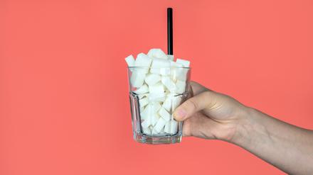 Eine junge Frau hält ein Glas mit Zuckerwürfeln und einem Strohhalm auf rosa Hintergrund (Symbolbild).