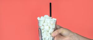 Eine junge Frau hält ein Glas mit Zuckerwürfeln und einem Strohhalm auf rosa Hintergrund (Symbolbild).