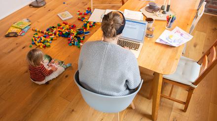 Ein zweijähriges Kind spielt im Wohnzimmer, während seine Mutter Zuhause im Homeoffice an einem Laptop arbeitet.