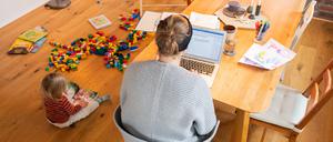 Ein zweijähriges Kind spielt im Wohnzimmer, während seine Mutter Zuhause im Homeoffice an einem Laptop arbeitet.