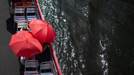 Touristen schützen sich auf einem Ausflugsschiff auf der Spree mit roten Sonnenschirmen. 