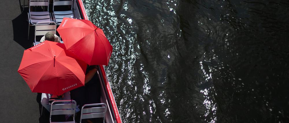 Touristen schützen sich auf einem Ausflugsschiff auf der Spree mit roten Sonnenschirmen.