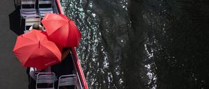 Touristen schützen sich auf einem Ausflugsschiff auf der Spree mit roten Sonnenschirmen.