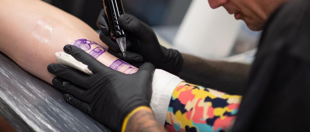Theodor Tinte tätowiert Hannah, Mitarbeiterin der BVG, den U-Bahnhof Neukölln auf ihr Bein.