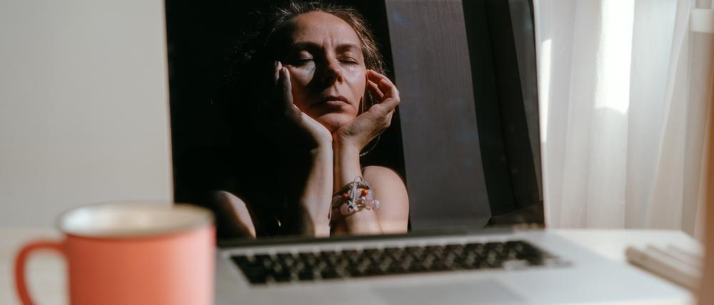 Eine Frau stützt den Kopf in die Hände, sie hält die Augen geschlossen, ihr Gesicht spiegelt sich im Bildschirm eines Laptops.