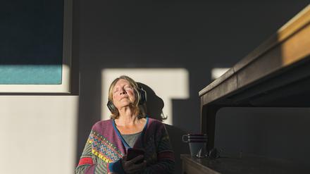 Eine ältere Frau sitzt auf einem Stuhl und hört mit Kopfhörern Musik.