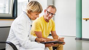 Ein mittelalter Mann sitzt auf einer Bank neben einer Ärztin, die ihm etwas auf einem Tablet zeigt.