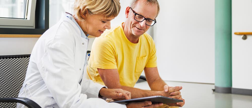Ein mittelalter Mann sitzt auf einer Bank neben einer Ärztin, die ihm etwas auf einem Tablet zeigt.