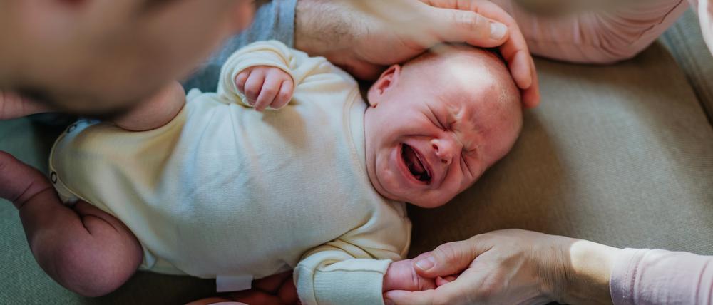Ein Baby in einem weißen Body liegt auf einer hellbraunen Unterlage, es schreit und wird dabei von den Händen seiner Eltern umfangen.