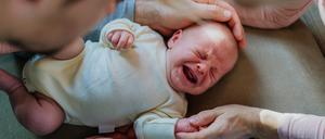 Ein Baby in einem weißen Body liegt auf einer hellbraunen Unterlage, es schreit und wird dabei von den Händen seiner Eltern umfangen.