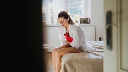 Eine junge Frau sitzt mit einer Wärmflasche auf einem Bett.