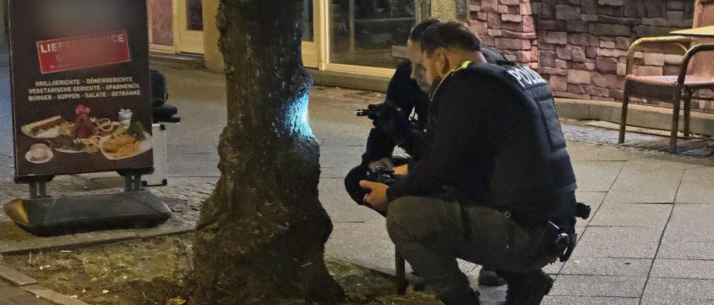Zwei Polizisten begutachten eine Spur, die eine Kugel an einem Baum hinterlassen hat. Ein Mann war durch einen Schuss ins Bein verletzt worden. Am Abend sei er an einem Imbiss in Reinickendorf angeschossen worden, teilte ein Sprecher der Polizei mit.
