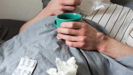 Ein Mann liegt mit Taschentüchern, Teetasse, Nasenspray und Tabletten im Bett (Symbolbild).
