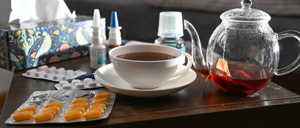 Medikamente und eine Kanne und Tasse Tee zur Bekämpfung einer Erkältung sind auf einem Nachttisch aufgestellt.