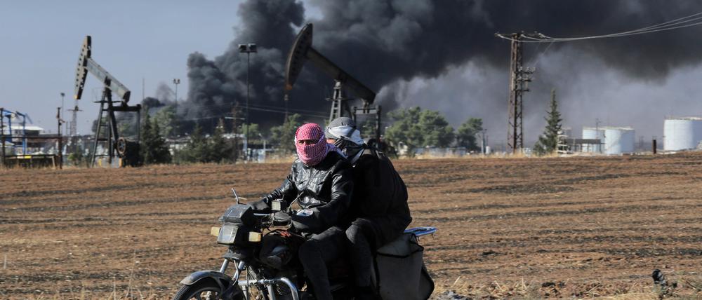 Menschen fahren auf einem Motorrad, während dichter Rauch aus einer Ölförderanlage aufsteigt, die am 25. Oktober 2024 nahe der nordöstlichen Grenze Syriens zur Türkei beschossen wurde (Archivbild).