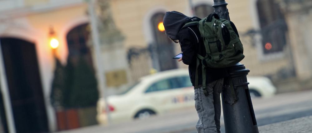 Ein Schulkind mit Schulrucksack steht angelehnt an einer Straßenlampe und ist mit einem elektronischen Spielgerät beschäftigt.
