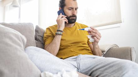 Ein Mann sitzt auf dem Sofa, in der einen Hand ein Fieberthermometer, in der anderen ein Handy am Ohr.
