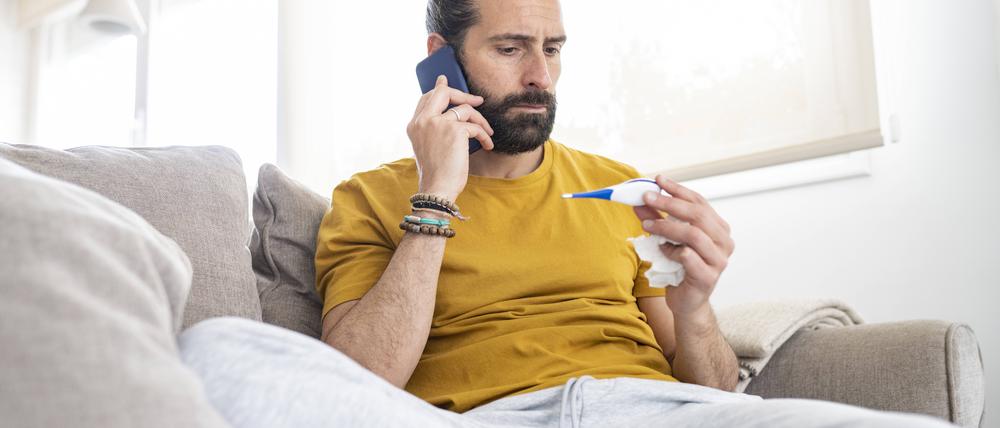 Ein Mann sitzt auf dem Sofa, in der einen Hand ein Fieberthermometer, in der anderen ein Handy am Ohr.