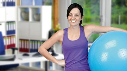 Eine Frau mit Medizinball unterm Arm lächelt frech in die Kamera, hinter ihr stehen Aktenordner.