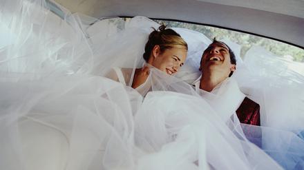 Ein lachendes Brautpaar umhüllt vom Kleid der Braut in einem Auto.