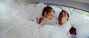 Ein lachendes Brautpaar umhüllt vom Kleid der Braut in einem Auto.