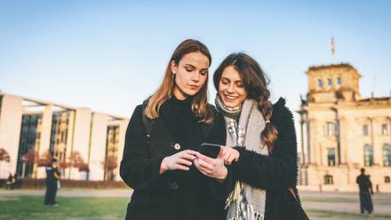 Handy im Mittelpunkt: Zwei junge Frauen vor dem Reichstag in Berlin schauen auf ein Smartphone. 