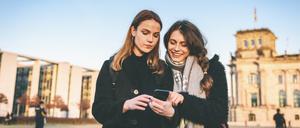 Handy im Mittelpunkt: Zwei junge Frauen vor dem Reichstag in Berlin schauen auf ein Smartphone. 